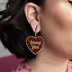May include: Red glitter heart-shaped earrings with gold trim and the text "Dump Him" in gold lettering.