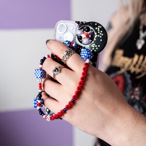 May include: A red and blue beaded phone charm with the words "Feed Me Pasta" spelled out. The charm is attached to a white phone case with a black and white moon design.