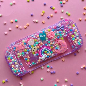 May include: A clear, purple Switch case adorned with colourful beads, stars, and charms. The case features a rainbow, a crescent moon, a rose, and the word "UGH". Star-shaped confetti is scattered around the case on a pink background.