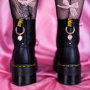May include: Black platform boots with yellow stitching and a gold heart charm. The boots have a black leather upper and a thick sole. The model is wearing black fishnet stockings with pink bows.