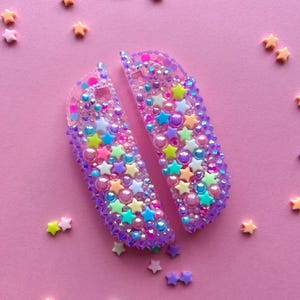 Puede incluir: Dos mandos Joy-Con Switch transparentes, con bordes morados, est&aacute;n decorados con cuentas en forma de estrella de colores, purpurina iridiscente y peque&ntilde;as cuentas redondas. Alrededor de los mandos hay sprinkles en forma de estrella en varios colores pastel, sobre un fondo rosa.