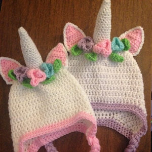 May include: Two handmade crochet unicorn hats. The hats are white with pink and purple accents. Each hat features a white horn, pink ears, and a floral crown with pink, purple, and blue flowers. Braided ties with tassels complete the design.