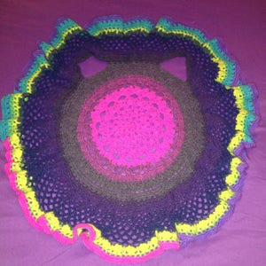 Op de afbeelding: Een handgemaakte, cirkelvormige gehaakte doily in een levendig kleurenpalet. Het midden is felroze, overgaand in bruin, paars en blauwgroen, met een gerimpelde rand in geel en roze. Het openwerkontwerp is zichtbaar.