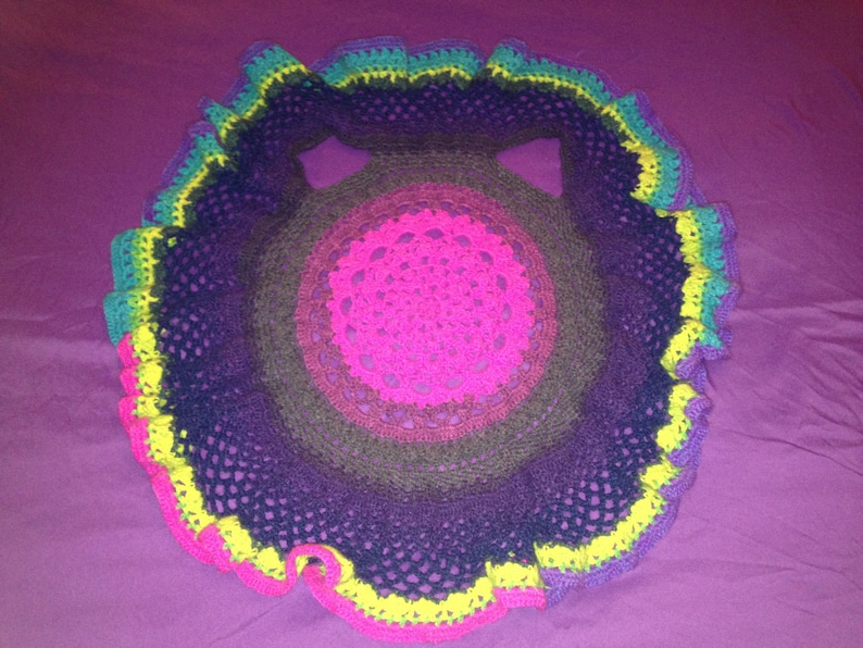 Op de afbeelding: Een handgemaakte, cirkelvormige gehaakte doily in een levendig kleurenpalet. Het midden is felroze, overgaand in bruin, paars en blauwgroen, met een gerimpelde rand in geel en roze. Het openwerkontwerp is zichtbaar.