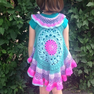May include: A handmade crochet vest in shades of teal, purple, and pink. The vest features a circular design in the center and a ruffled hem. The vest is worn over a teal t-shirt.