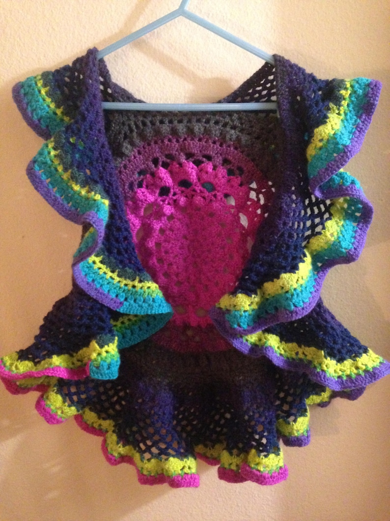 Op de afbeelding: Een handgemaakt gehaakt vest in tinten paars, teal, geel en roze. Het vest heeft een cirkelvormig ontwerp in het midden en gerimpelde randen. Het kledingstuk wordt op een blauwe hanger getoond.