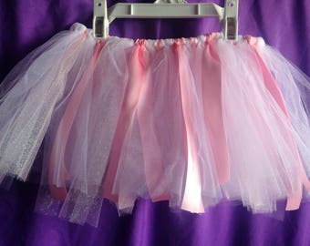 Op de afbeelding: Een roze en witte tule tutu rok met roze satijnen linten die aan de tailleband hangen. De rok hangt aan een witte hanger tegen een paarse achtergrond. De tule heeft een sprankelend, glinsterend uiterlijk.