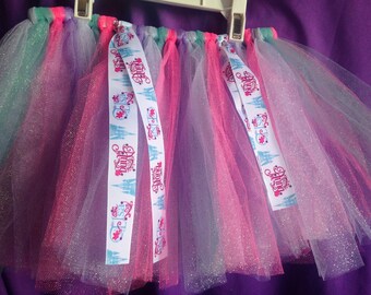 Op de afbeelding: Een kleurrijke tutu rok met lagen roze, paars en turquoise tule. De rok is versierd met witte linten met de naam "Anna" en een kasteelontwerp. De tule heeft een glitterafwerking, wat de rok extra glans geeft.
