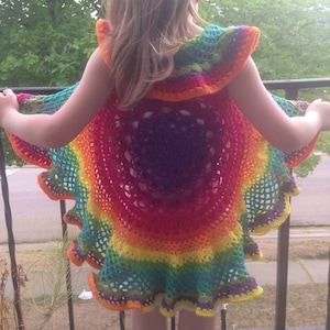 Op de afbeelding: Een levendige, regenboogkleurige gehaakte cape met een gerimpelde rand. De cape heeft een cirkelvormig ontwerp met een kleurengradatie, waaronder rood, oranje, geel, groen en paars. Het kind staat op een balkon, met bomen op de achtergrond.