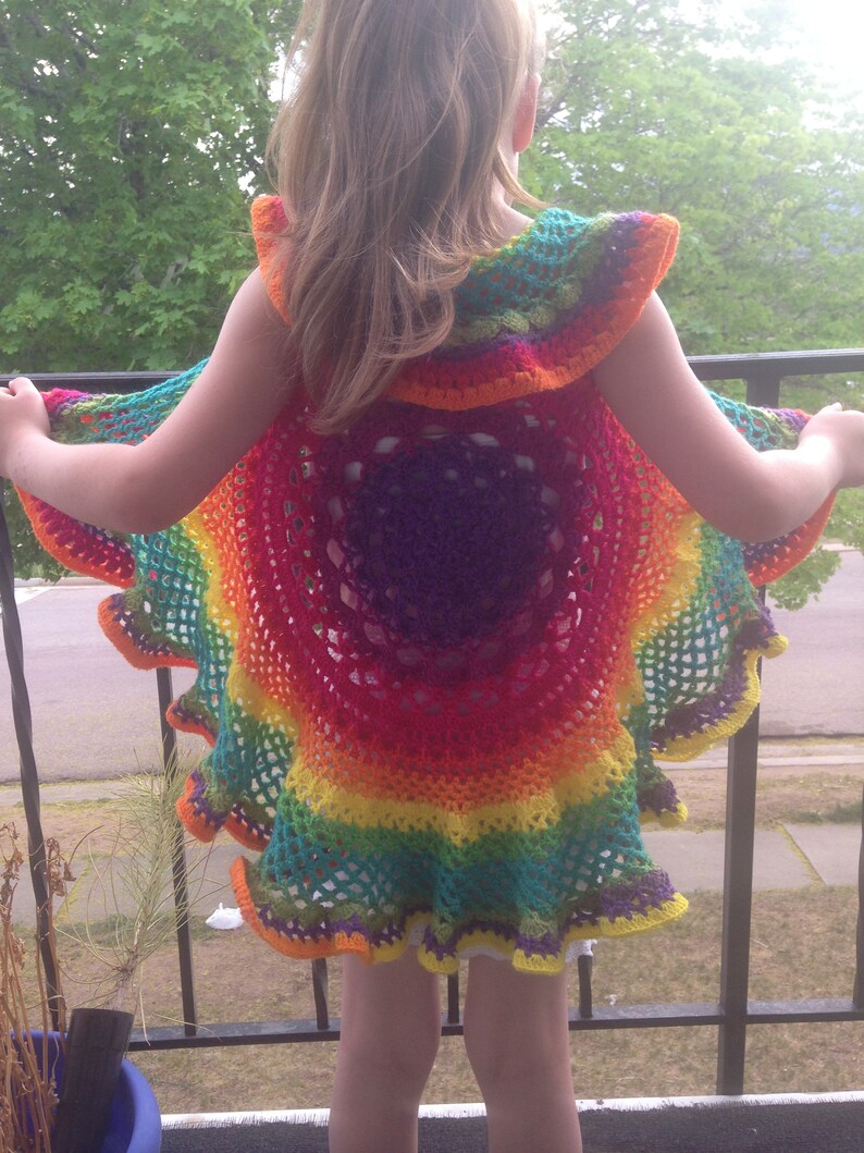 Op de afbeelding: Een levendige, regenboogkleurige gehaakte cape met een gerimpelde rand. De cape heeft een cirkelvormig ontwerp met een kleurengradatie, waaronder rood, oranje, geel, groen en paars. Het kind staat op een balkon, met bomen op de achtergrond.