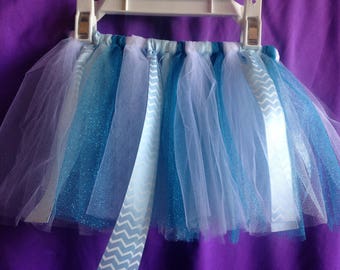 Op de afbeelding: Een blauw-witte tutu rok met lagen tule en lint. De rok heeft blauwtinten, waaronder een glinsterende stof en wit zigzagpatroon lint. De rok hangt aan een witte hanger tegen een paarse achtergrond.