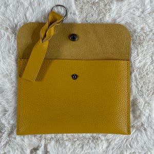 May include: Mustard yellow leather wallet with a silver key ring and a braided leather strap. The wallet has a snap closure and a textured surface. The interior is a lighter shade of yellow.