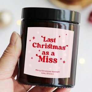 May include: A dark brown glass jar with a black lid, held in a hand. The jar features a pink label with red text that reads "Last Christmas as a Miss." The label also includes the text "Merry Christmas Hannah! Love, Olivia x."