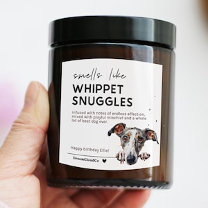 May include: A brown glass candle jar with a black lid. The label reads "smells like WHIPPET SNUGGLES" with a Whippet dog illustration. The text also includes "Happy birthday Ellie!" and "DreamCloudCo".