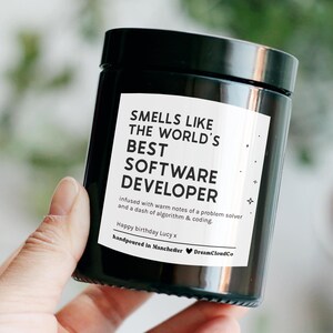 May include: A black candle jar with a white label that reads "Smells like the world's best software developer" and "infused with warm notes of a problem solver and a dash of algorithm & coding". The label also includes the text "Happy birthday Lucy x" and "handpoured in Manchester DreamCloudCo".