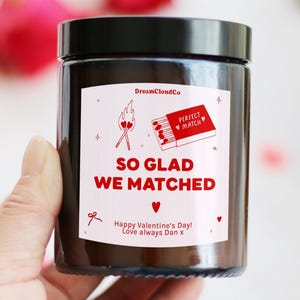 May include: A brown glass jar with a black lid, displaying a label with red text and illustrations. The label says "SO GLAD WE MATCHED" and "Happy Valentine's Day! Love always Dan x". The jar is held by a hand, with red roses in the background.