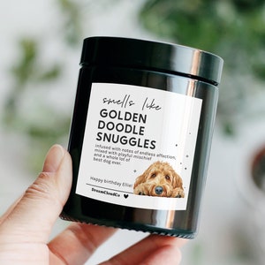 May include: A black candle jar with a white label that reads "smells like GOLDEN DOODLE SNUGGLES" and features a cartoon illustration of a golden doodle dog. The label also includes the text "infused with notes of endless affection, mixed with playful mischief and a whole lot of best dog ever." The label is signed "DreamCloudCo" and includes the text "Happy birthday Elliet".
