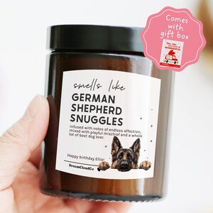 May include: A brown glass candle jar with a black lid. The label reads "smells like GERMAN SHEPHERD SNUGGLES." A pink gift box sticker is attached. The candle is held in a hand.