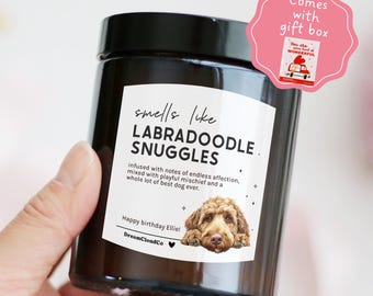 May include: A black jar candle with a white label that reads "smells like LABRADOODLE SNUGGLES." The label also includes the text "infused with notes of endless affection" and a picture of a Labradoodle. A pink gift box is attached.