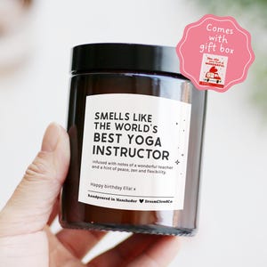 May include: A dark brown glass candle with a black lid. The label says "Smells Like The World's Best Yoga Instructor." A pink gift box is attached. The candle is hand-poured in Manchester.