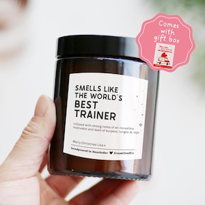 May include: A brown glass candle with a black lid. The label says "SMELLS LIKE THE WORLD'S BEST TRAINER." The candle is hand-poured in Manchester. A pink gift box sticker is attached. The candle is held in a hand.