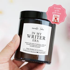 May include: A dark glass jar candle with a black lid and a white label. The label reads "smells like IN MY WRITER ERA" with additional text. A pink gift box sticker is in the upper right corner.