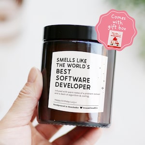 May include: A dark brown glass candle with a black lid and a white label. The label says "Smells Like The World's Best Software Developer." A pink gift box tag is attached. The candle is hand-poured in Manchester.