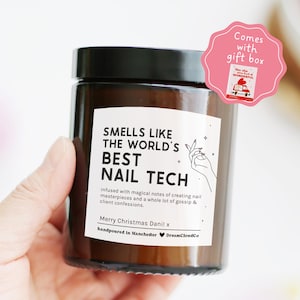 May include: A brown glass jar with a black lid, labelled "SMELLS LIKE THE WORLD'S BEST NAIL TECH." The label includes text about nail masterpieces and gossip. A pink gift box sticker reads "Comes with gift box."