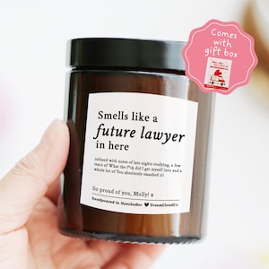 May include: A dark brown candle jar with a black lid, featuring a label that reads "Smells like a future lawyer in here." The label includes a message and is handpoured in Manchester. A pink gift box sticker is attached.