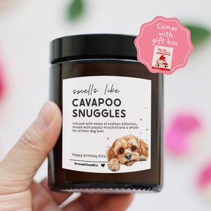 May include: A brown glass candle jar with a black lid. The label reads "smells like CAVAPOO SNUGGLES" with a dog illustration. A pink gift box tag is attached. The candle is held in a hand.