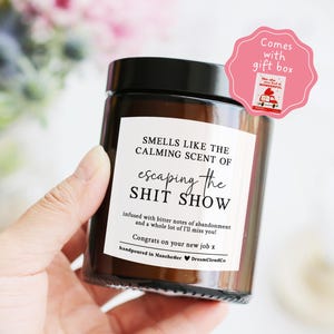 May include: A brown glass candle with a black lid, labelled "SMELLS LIKE THE CALMING SCENT OF escaping the SHIT SHOW." The candle is hand-poured in Manchester and comes with a gift box, perfect for a new job.