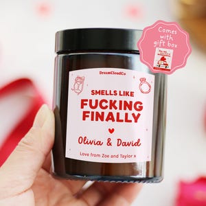 May include: A brown glass jar candle with a black lid. The label reads "SMELLS LIKE FUCKING FINALLY" in red text, with the names "Olivia & David" below. A pink gift box is included.