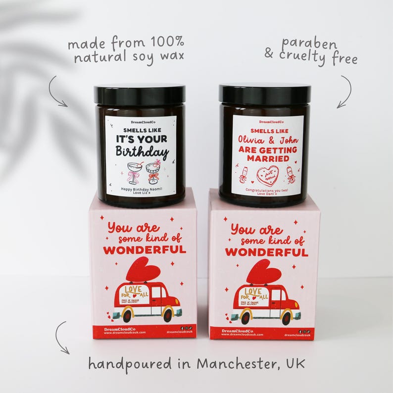 May include: Two dark brown glass candles with black lids, each on a pink box. The candles have labels that read "Smells Like It's Your Birthday" and "Smells Like Olivia & John Are Getting Married." The boxes say "You are some kind of Wonderful."