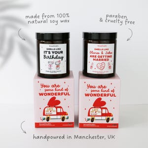 Puede incluir: Dos velas de vidrio oscuro con tapas negras, etiquetadas como "Smells Like It's Your Birthday" y "Smells Like Olivia & John Are Getting Married." Cada vela se encuentra sobre una caja rosa con un gr&aacute;fico de coche rojo y el texto "You are some kind of WONDERFUL."