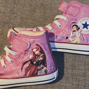 K&ouml;nnte beinhalten: Ein Paar kn&ouml;chelhohe Converse-Sneaker in schimmerndem Pink. Jeder Schuh verf&uuml;gt &uuml;ber wei&szlig;e Schn&uuml;rsenkel, eine wei&szlig;e Gummikappe und eine wei&szlig;e Sohle mit schwarzen Streifen. Die Seiten zeigen farbenfrohe Illustrationen von Anime-Charakteren.
