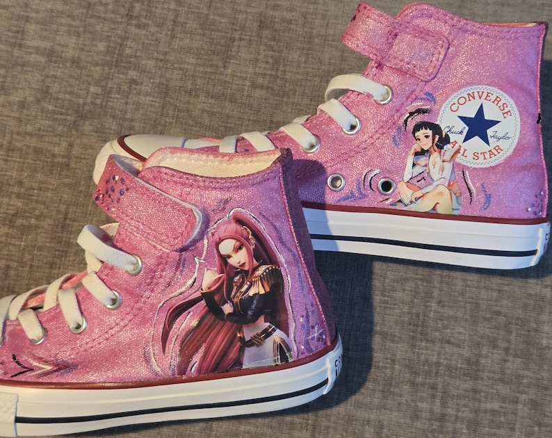 K&ouml;nnte beinhalten: Ein Paar kn&ouml;chelhohe Converse-Sneaker in schimmerndem Pink. Jeder Schuh verf&uuml;gt &uuml;ber wei&szlig;e Schn&uuml;rsenkel, eine wei&szlig;e Gummikappe und eine wei&szlig;e Sohle mit schwarzen Streifen. Die Seiten zeigen farbenfrohe Illustrationen von Anime-Charakteren.