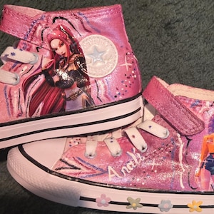 K&ouml;nnte beinhalten: Ein Paar rosa High-Top-Sneaker mit handgemalten Anime-Figuren an den Seiten. Die Schuhe haben wei&szlig;e Schn&uuml;rsenkel, einen Klettverschluss, eine wei&szlig;e Gummikappe und eine wei&szlig;e Zwischensohle, die mit kleinen bunten Blumenakzenten verziert ist.