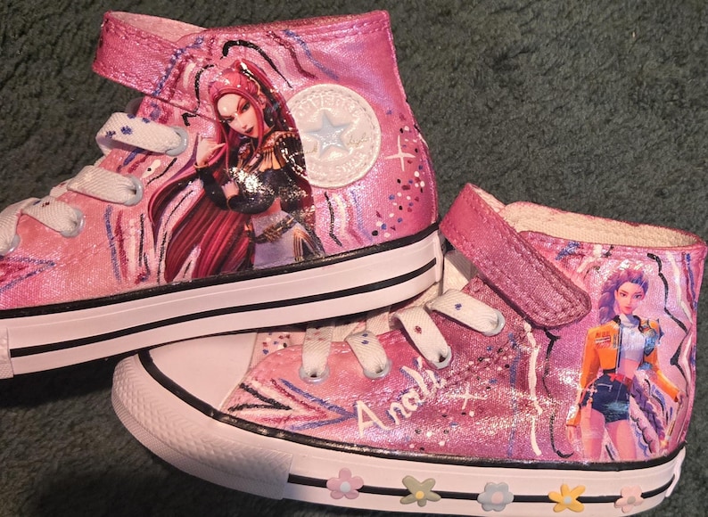 K&ouml;nnte beinhalten: Ein Paar rosa High-Top-Sneaker mit handgemalten Anime-Figuren an den Seiten. Die Schuhe haben wei&szlig;e Schn&uuml;rsenkel, einen Klettverschluss, eine wei&szlig;e Gummikappe und eine wei&szlig;e Zwischensohle, die mit kleinen bunten Blumenakzenten verziert ist.