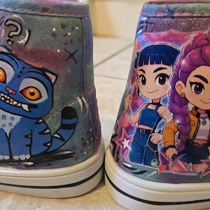 K&ouml;nnte beinhalten: Ein Paar High-Top-Sneaker mit glitzerndem violett-blauem Galaxie-Hintergrund. Der linke Schuh zeigt eine blaue Katze mit orangefarbenen Augen und Fragezeichen, w&auml;hrend der rechte Schuh drei Zeichentrickfiguren mit buntem Haar darstellt.