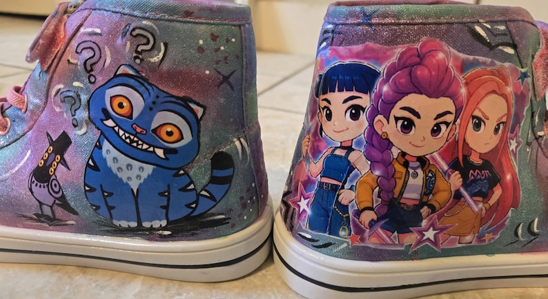K&ouml;nnte beinhalten: Ein Paar High-Top-Sneaker mit glitzerndem violett-blauem Galaxie-Hintergrund. Der linke Schuh zeigt eine blaue Katze mit orangefarbenen Augen und Fragezeichen, w&auml;hrend der rechte Schuh drei Zeichentrickfiguren mit buntem Haar darstellt.