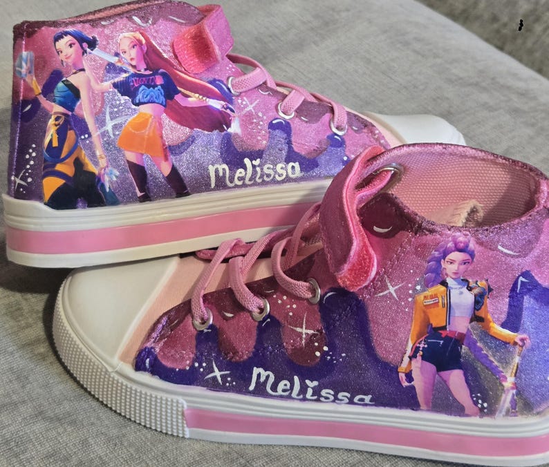 Może przedstawiać: Para wysokich trampek w odcieniach r&oacute;żu i fioletu z brokatowym wykończeniem. Buty posiadają nadrukowane postacie w stylu anime, białe gumowe noski, r&oacute;żowe sznurowadła oraz zapięcie na rzep. Imię Melissa jest namalowane białą czcionką.