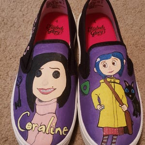 May include: Pair of custom-painted slip-on shoes featuring characters from the movie "Coraline." One shoe depicts Coraline with the text "Coraline," and the other shows Coraline in a yellow raincoat with a black cat. The shoes have purple uppers and white soles.