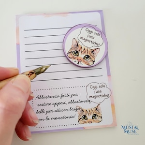 以下が含まれることがあります： 猫をテーマにしたマグネットが付いた、罫線入りのメモ帳。マグネットには、吹き出しの中に「Oggi solo fusa magnetiche!」というテキストが入った猫のイラストが描かれています。イタリア語のテキストも追加されています。