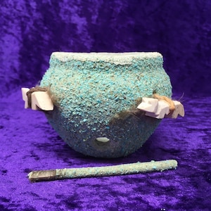 May include: A small, round, turquoise-colored ceramic pot with two white bone-like pieces attached to the sides. The pot has a small hole in the bottom and a wooden stick is lying next to it.