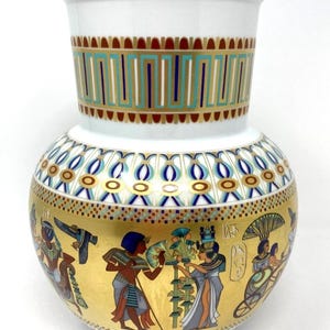 May include: A white ceramic vase with gold trim and an Egyptian-themed design. The vase features a gold band with hieroglyphics and figures, and a geometric pattern in teal, gold, and navy blue.
