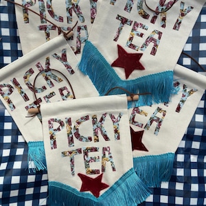 Könnte beinhalten: Vier weiße Stoffbanner mit blauen Fransen und einem roten Stern. Jedes Banner sagt "Picky Tea" in floralen Buchstaben.