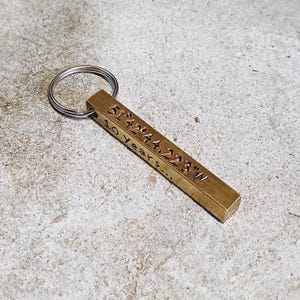 May include: A brass keychain with a silver key ring. The keychain is engraved with the coordinates "51.5044.2233" and the text "10 years".
