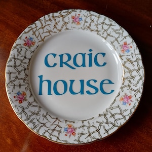 May include: White ceramic plate with a gold rim and a decorative pattern. The plate features the words "craic house" in teal blue. Small floral designs are placed around the rim.