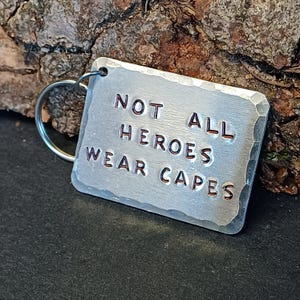 May include: Silver-toned keychain with a rectangular tag that reads "Not All Heroes Wear Capes".