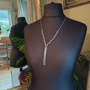 Può includere: Una collana color argento con un design a forma di Y. La collana presenta una delicata catena che si divide in due fili adornati con piccoli abbellimenti raggruppati. Una lunga frangia di catena pende dal basso. La collana è esposta su un manichino nero.